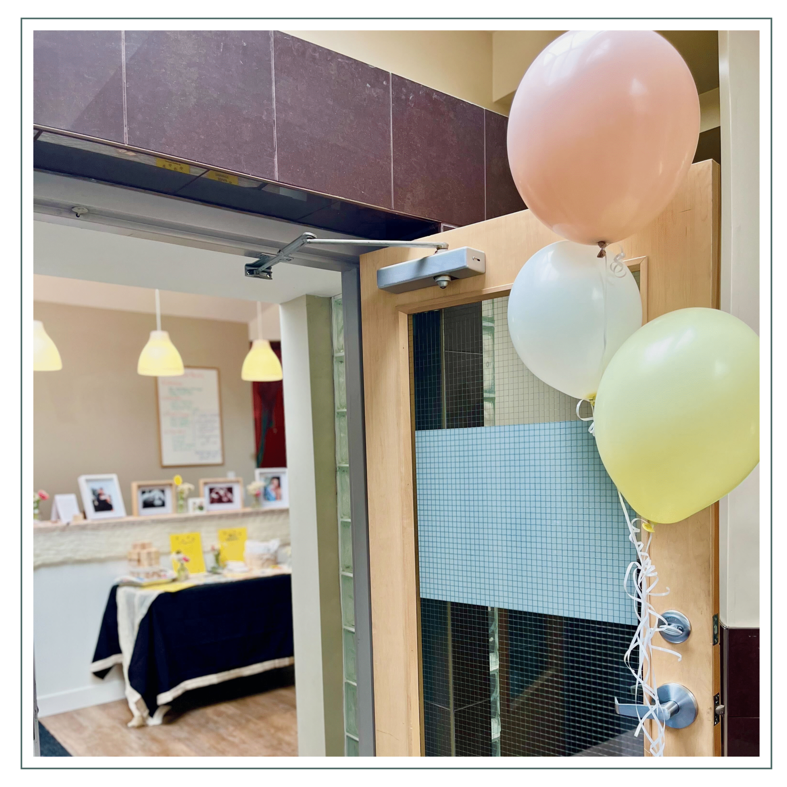 The image shows a welcoming entrance to a room with colorful balloons tied to the door. Inside, a table with photos and a few decorations suggests a celebratory or special event taking place. The space has a bright, inviting feel.