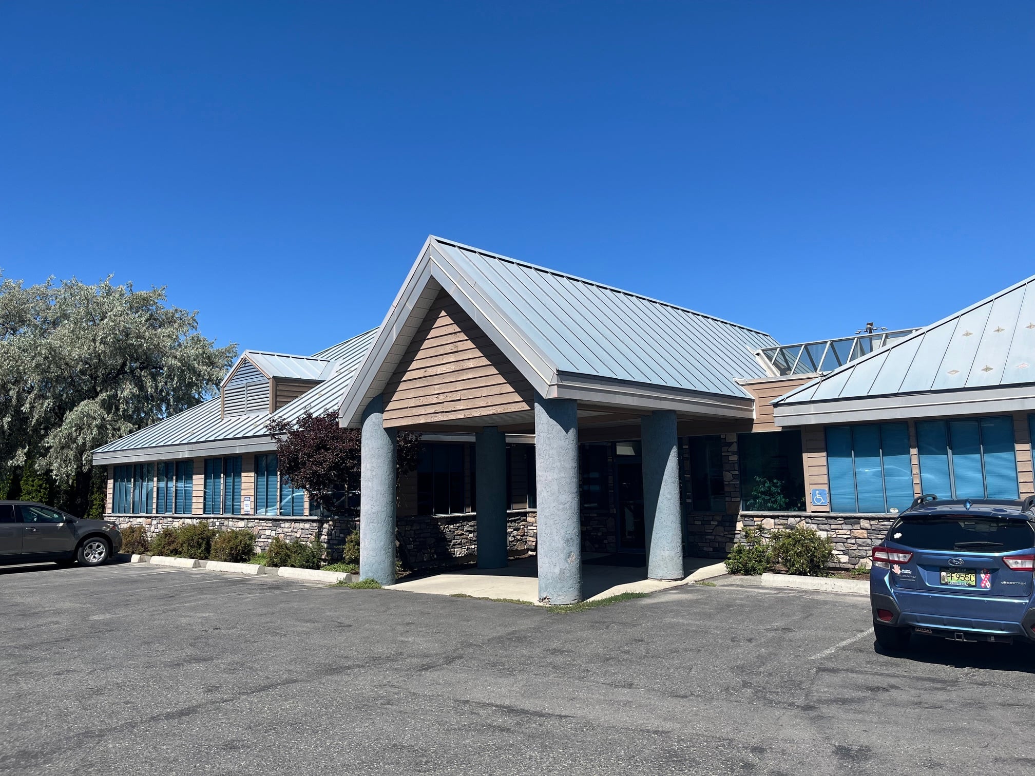 It's a picture of a building with a metal roof, stone walls, and a parking lot in front. The sky is clear.
