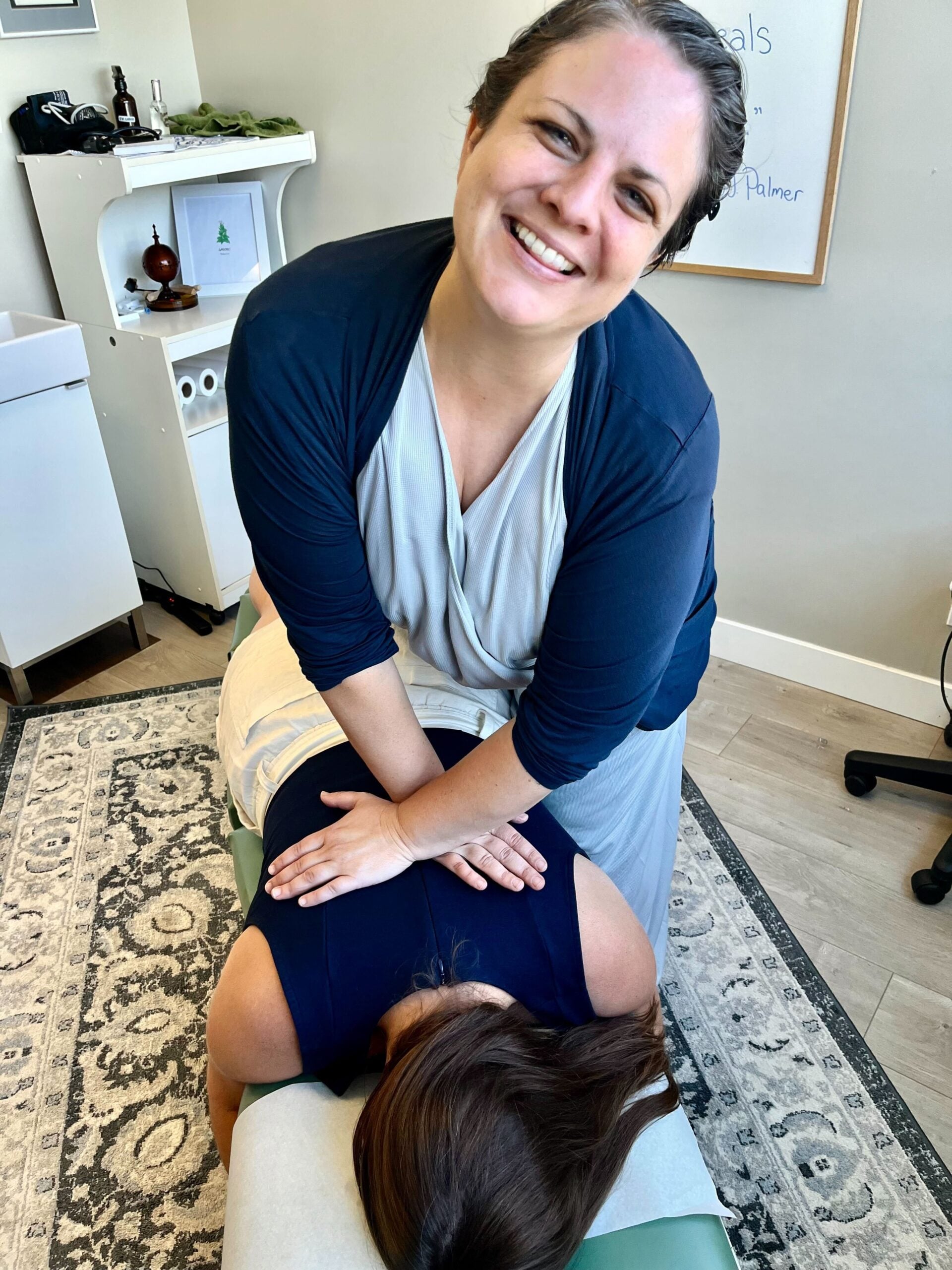 A woman is smiling while performing a chiropractic adjustment on a patient lying face down on a treatment table. The room has a cozy feel with a rug and minimal decor.