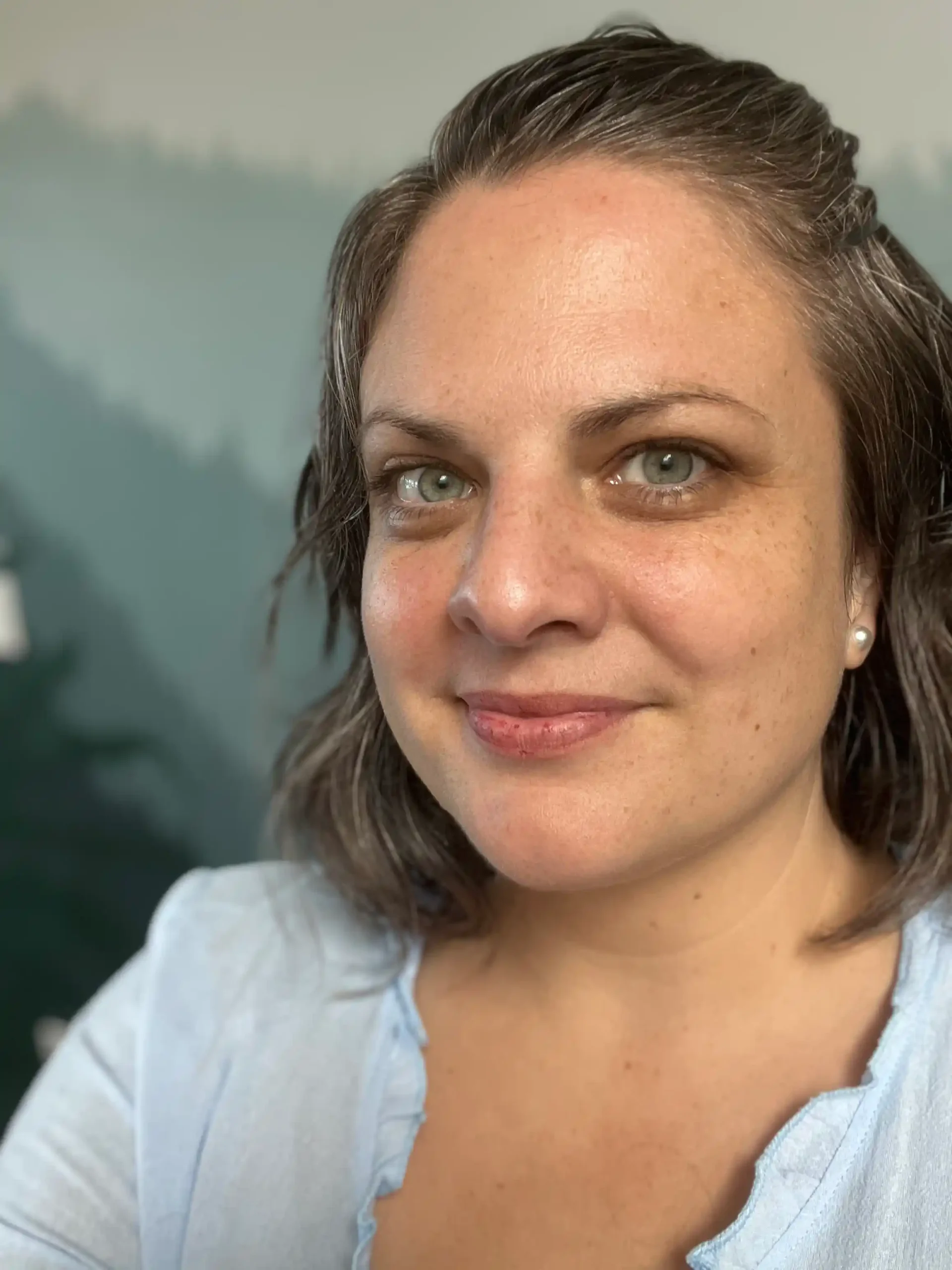 A woman is smiling softly in a close-up shot. She has short, slightly wavy hair and is wearing a light blue shirt. The background has a blurred green landscape.